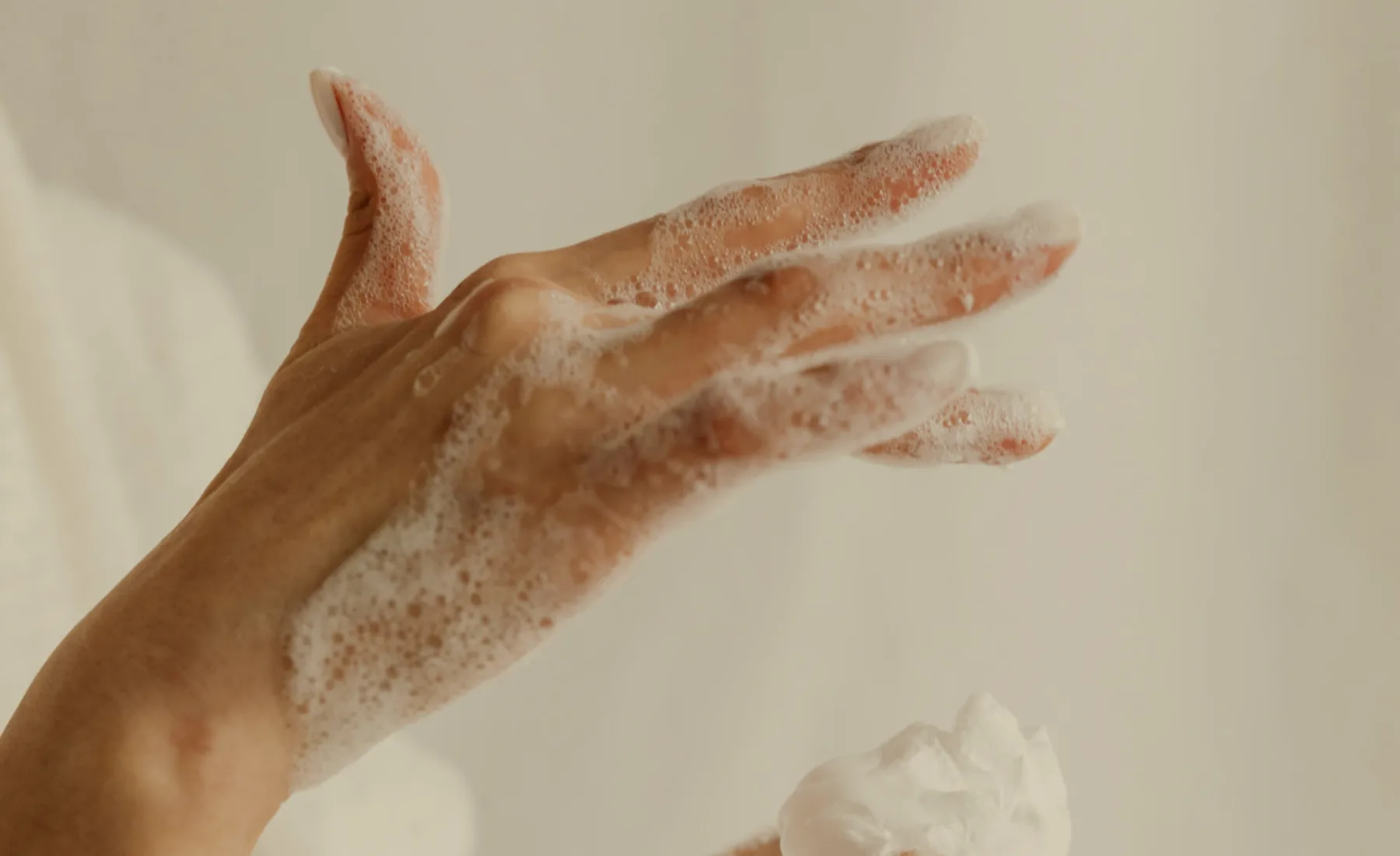 Manos aplicando espuma limpiadora sobre fondo crema cálido.